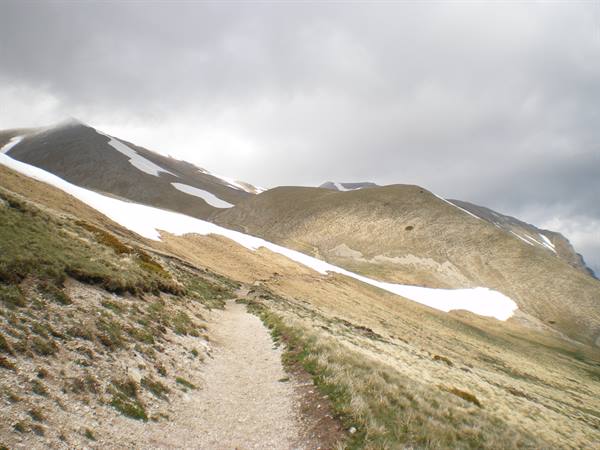 Gravel trail through dry grass and leftover snow, mountains under grey clouds