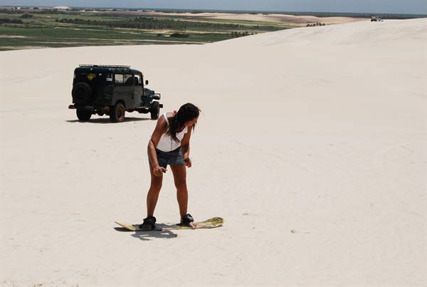 Girl tries sandboarding on white sand dune, black jeep parked nearby