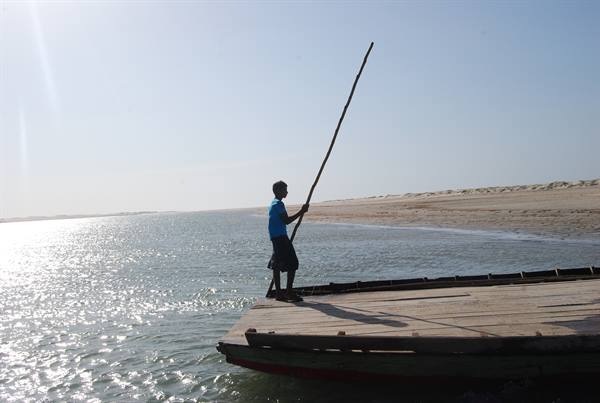 Boy with long pole steers a wooden ferry toward the sandy shore