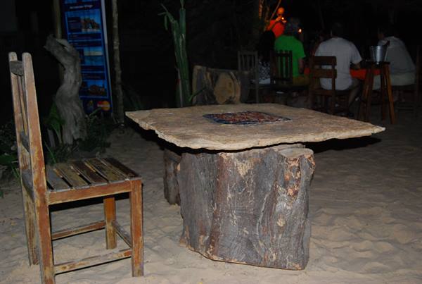 Stone table on a trunk, wooden chair, sand and people sitting at night