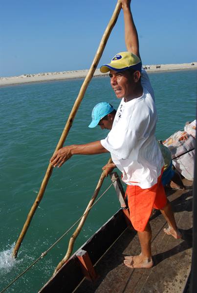 Man in yellow cap pushes a long wooden pole from the boat