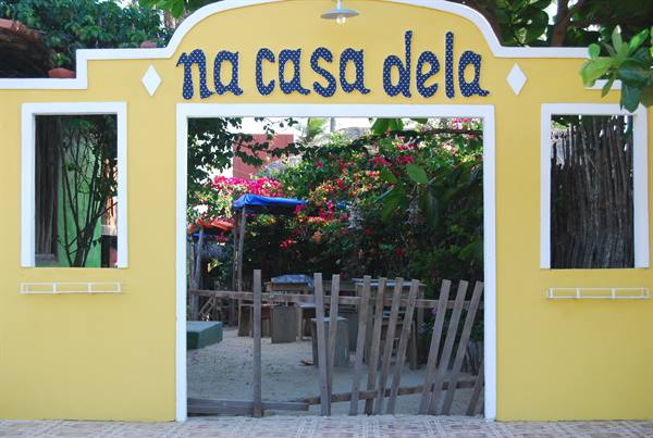 Yellow entrance with blue "Na Casa Dela" sign and wooden gate