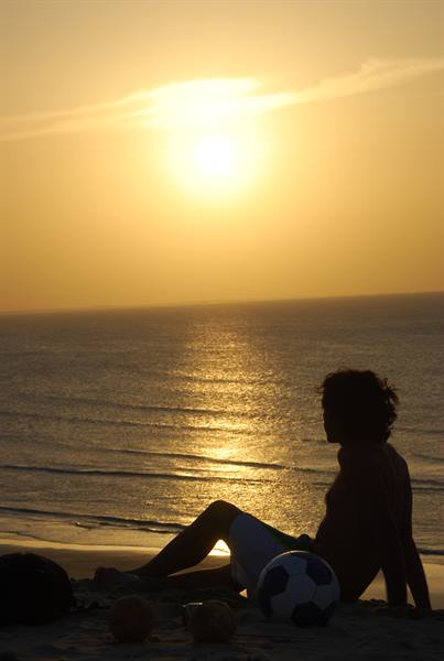 Boy sitting on the sand beside a football, facing the low sun over the sea