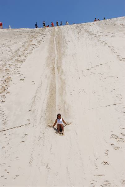 Girl slides down white sand dune, people at the top under blue sky