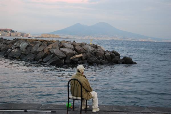 Fisherman on a chair by the breakwater rocks, Mount Vesuvius rising over Naples