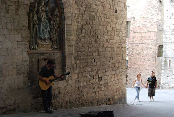 Guitarist plays beneath a painted shrine, stone walls, couple strolls by