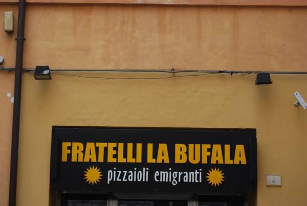 Black sign with yellow lettering Fratelli La Bufala pizzaioli emigranti on ochre wall