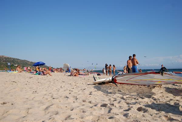 Sandy beach with sunbathers, blue umbrella and windsurf boards in foreground