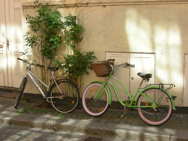 Green bicycle with wicker basket and a grey one leaning against a pale wall