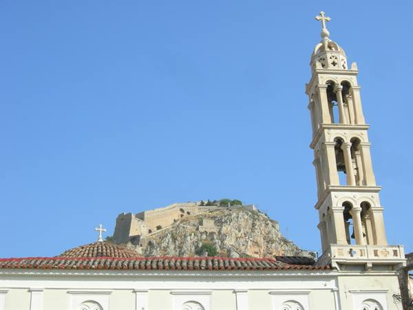 Stone bell tower beside white church, Palamidi fortress in the background