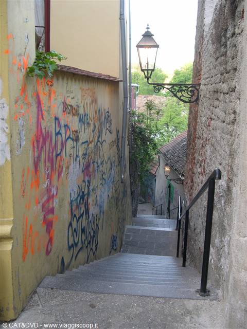 Narrow staircase with graffiti wall and wrought-iron lamp, descending between old houses
