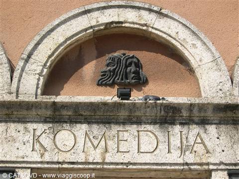 Bronze theatrical mask above the KOMEDIJA inscription on pale stone