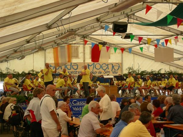 Banda Moena playing under a tent, musicians in yellow and crowd seated at tables