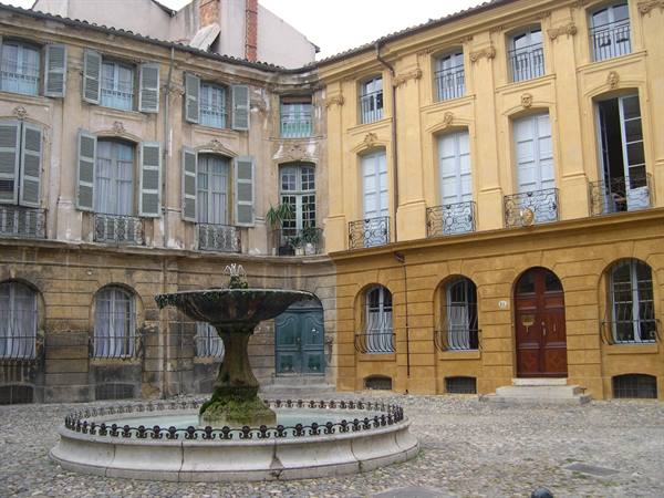 Stone fountain on cobbles, ochre facades with shutters in Aix-en-Provence