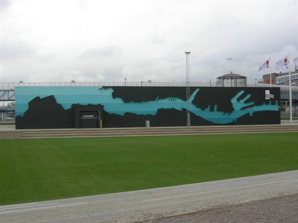 Wall painted with harbor map, green lawn in front, cloudy sky