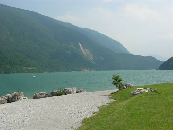 Molveno lake, gravel shore with rocks, green grass and forested mountains