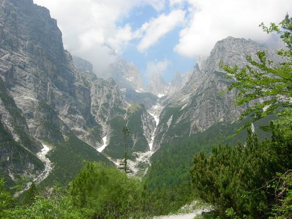 Rocky Dolomite gorge with residual snow, green conifers and clouds over the peaks