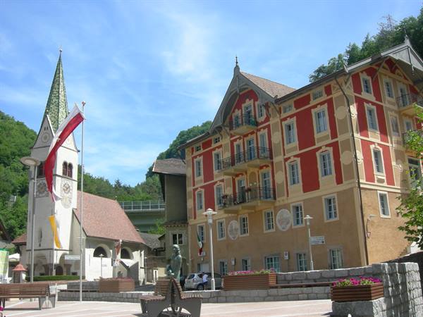 Whitewashed bell tower with green spire beside red frescoed building, sunny town square