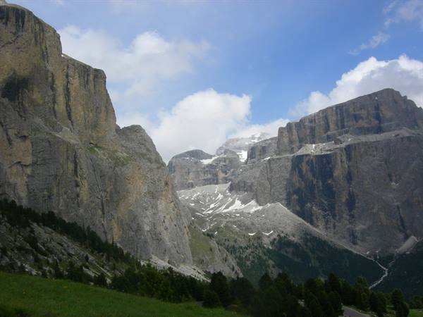 Dolomite cliffs above green meadow, patches of snow on rock, road below