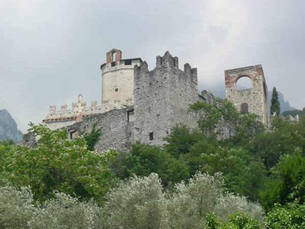 Stone castle with battlements and towers above olive trees, cloudy sky