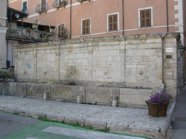 Old stone fountain with long basins, in front of pink buildings