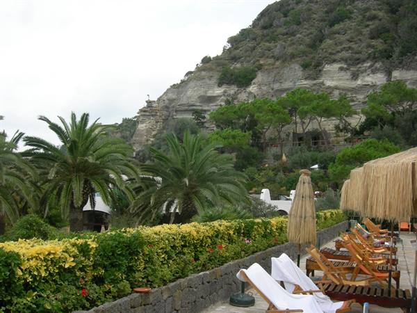 Wooden loungers and straw umbrellas among palms below a green tuff cliff