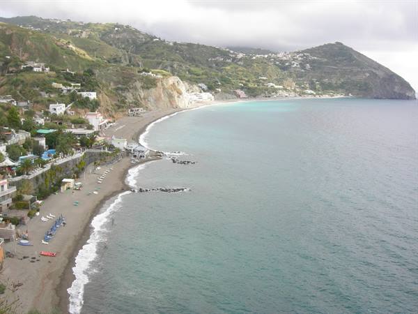 Dark sand beach beneath green hills, white houses and umbrellas along the shore