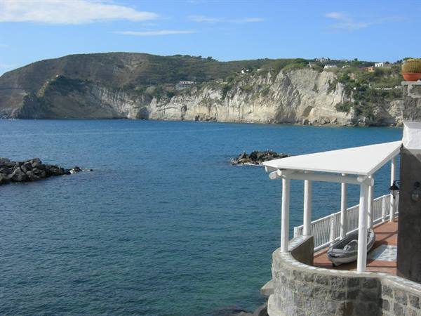 White pergola terrace with small boat, facing tuff cliffs and turquoise bay, Ischia