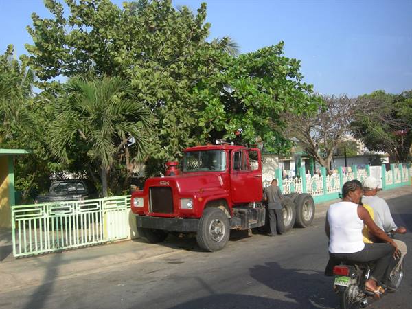 Red Mack truck on the road, men riding a motorcycle, green fences and palms