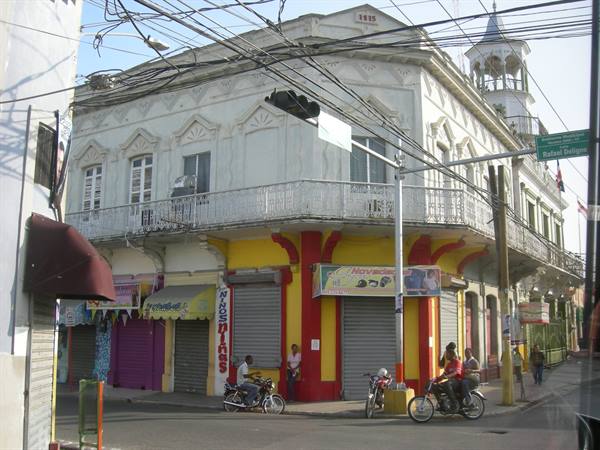 White colonial building with wrought-iron balcony, colorful shops, motorcyclists at the intersection