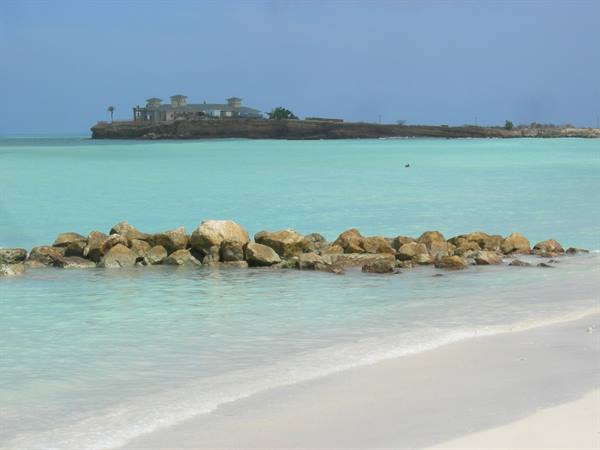Rocks on turquoise water by a white sand beach, buildings on an islet behind