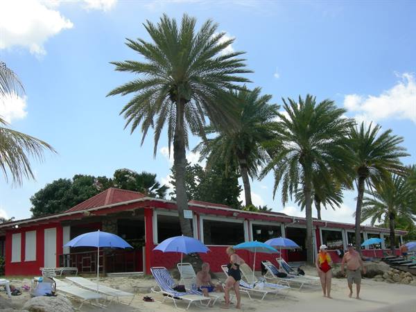 Tourists under blue umbrellas near a red beach bar among tall palm trees