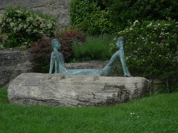 Green bronze sculpture of two reclining figures on boulder, flowering garden