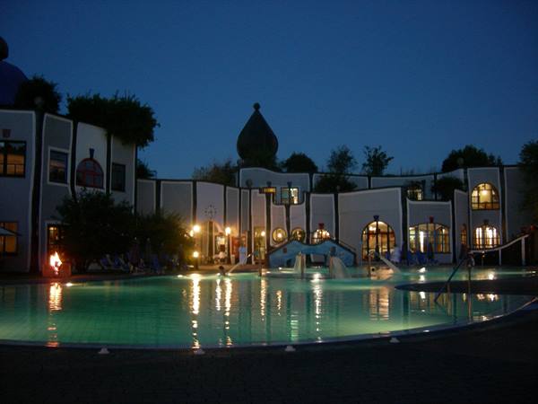 Illuminated thermal pool at night in front of colorful Hundertwasser buildings, Rogner Bad Blumau