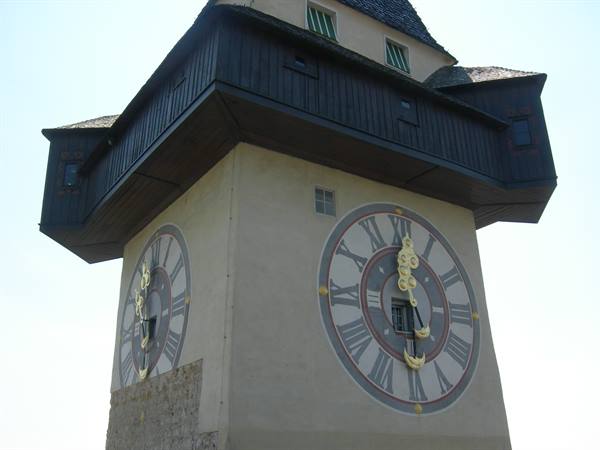 Graz clock tower, painted dials with golden hands, dark wooden balcony, blue sky