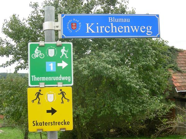 Tourist signs on metal pole in Blumau, soft daylight, trees and tiled roof behind