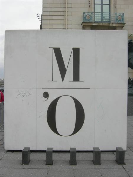White marble panel with black M'O letters, in front of stone building