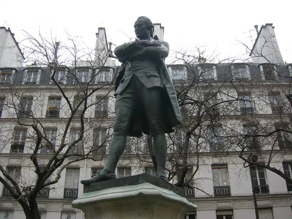 Bronze statue of a man in coat, arms crossed, in front of a Haussmann building