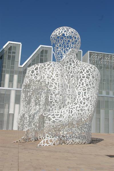 White sculpture of a seated figure made of interwoven metal letters, before glass buildings