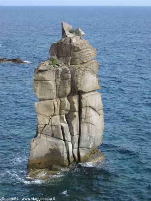 Tall solitary granite sea stack rising from deep blue water, cracked and vegetated