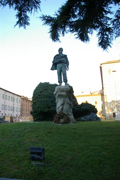 Bronze partisan statue on stone base, lawn and buildings around