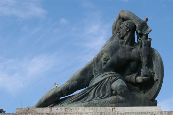 Bronze statue of muscular reclining giant with shield against blue sky