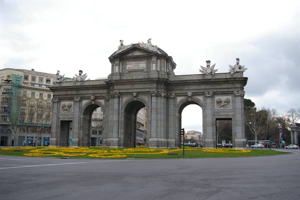 Puerta de Alcalá in Madrid, granite arches under cloudy sky, yellow flowerbed