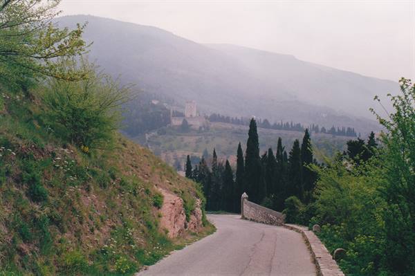 Asphalt road winding past cypress trees, hazy hills and a distant castle