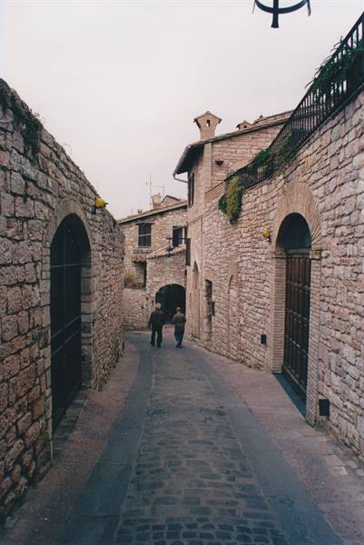 Stone alley with two people walking toward arches and medieval houses