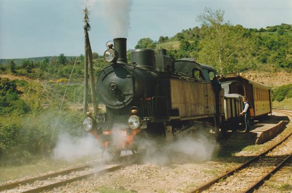Black steam locomotive releasing steam on tracks, driver standing beside carriage in countryside
