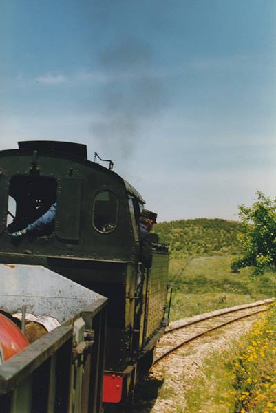 Green steam locomotive with driver at the window, smoke over curving track among hills