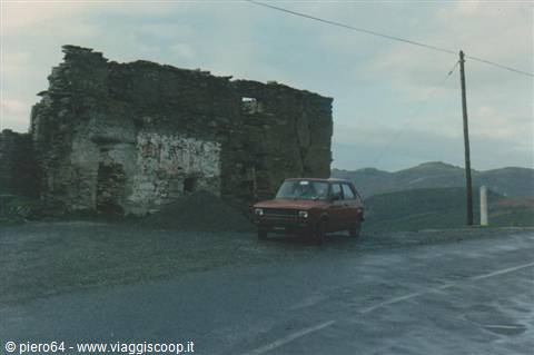 Red car parked on wet road beside old stone ruins among hills