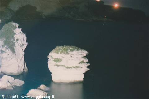 White sea stacks rise from dark water, distant lights on the cliff