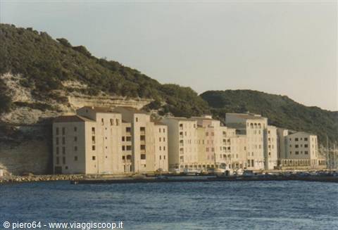 Pale buildings along Bonifacio harbor beneath the green hill